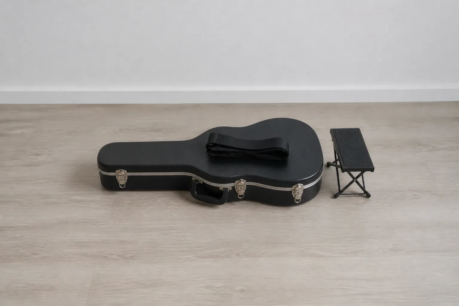Closed guitar case with a folded strap beside a low footstool on a neutral studio floor